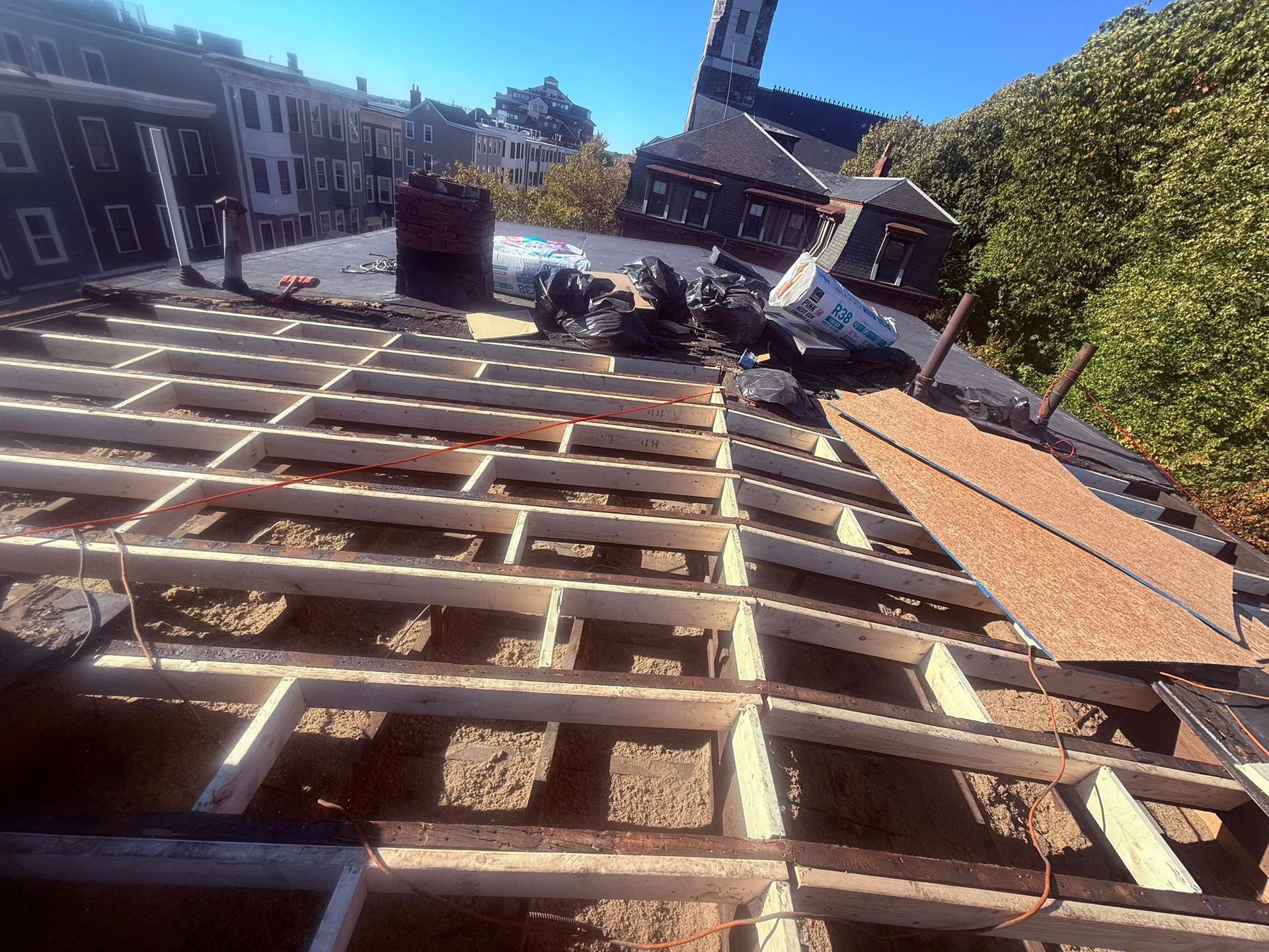 Roof under construction, showing exposed rafters, debris, and cityscape background.