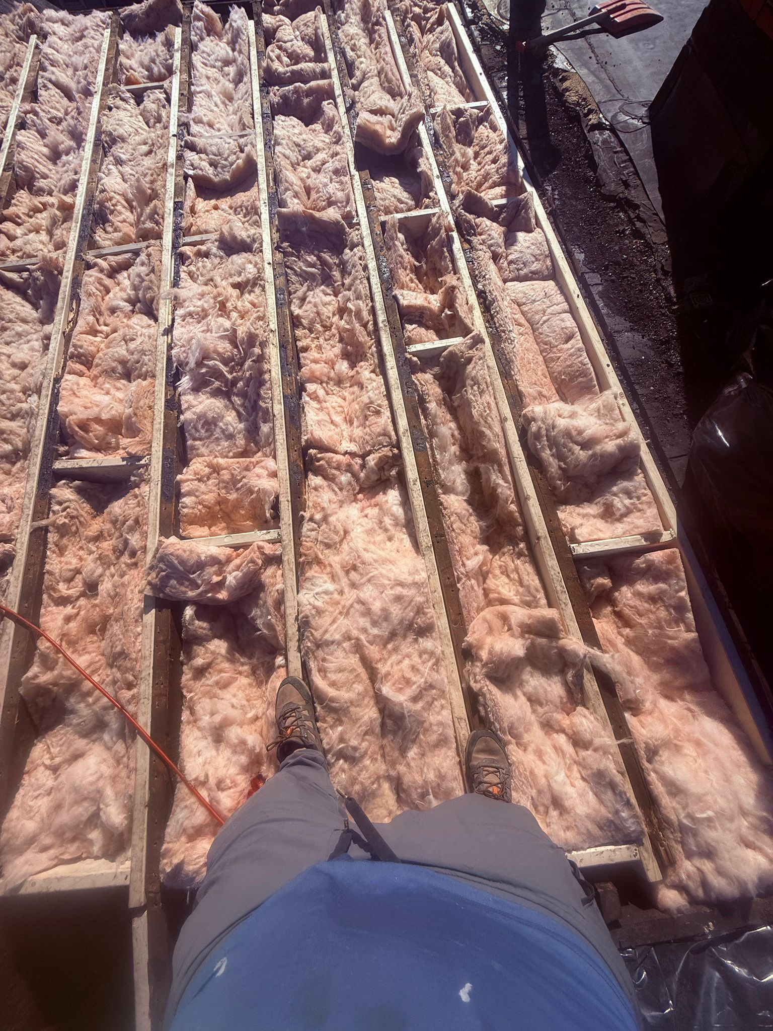 Person standing on a roof with exposed insulation between wooden beams.