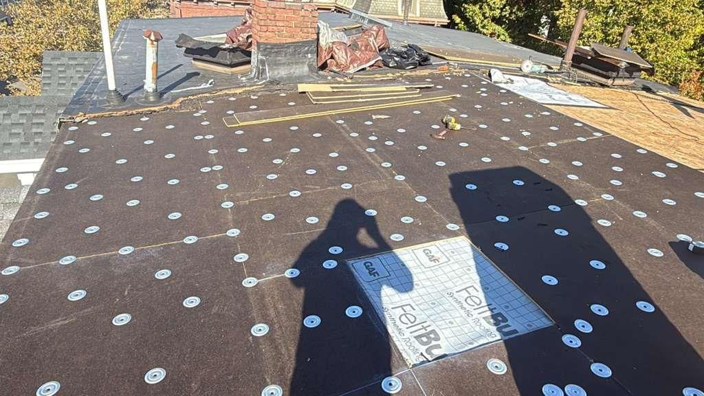 Flat roof under construction; dark asphalt with rows of fasteners, chimney, sunny outdoors.