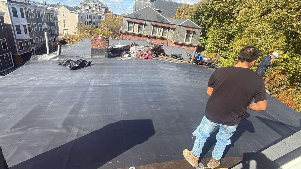 Workers on a flat roof install a dark membrane. City buildings and trees are in the background.