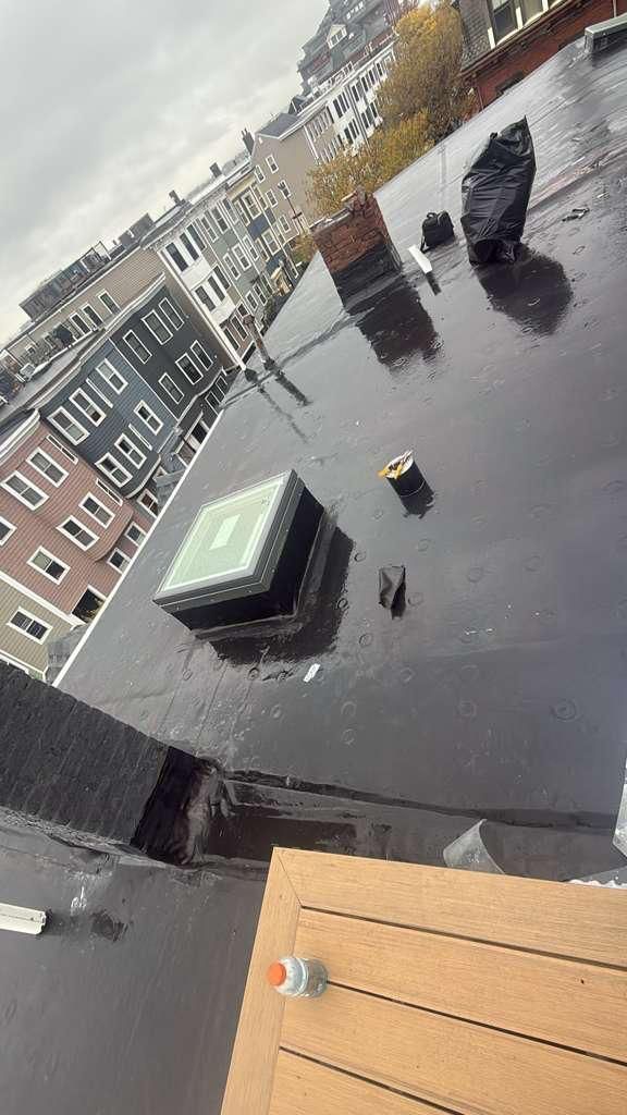 A wet, flat rooftop with a skylight, vent, and city buildings in the background. Overcast sky.