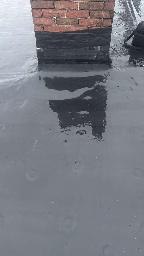 A brick chimney on a dark, wet roof surface reflecting its form.