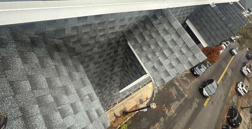 Overhead view of a building's shingled roof, a street with parked cars, and a sidewalk.