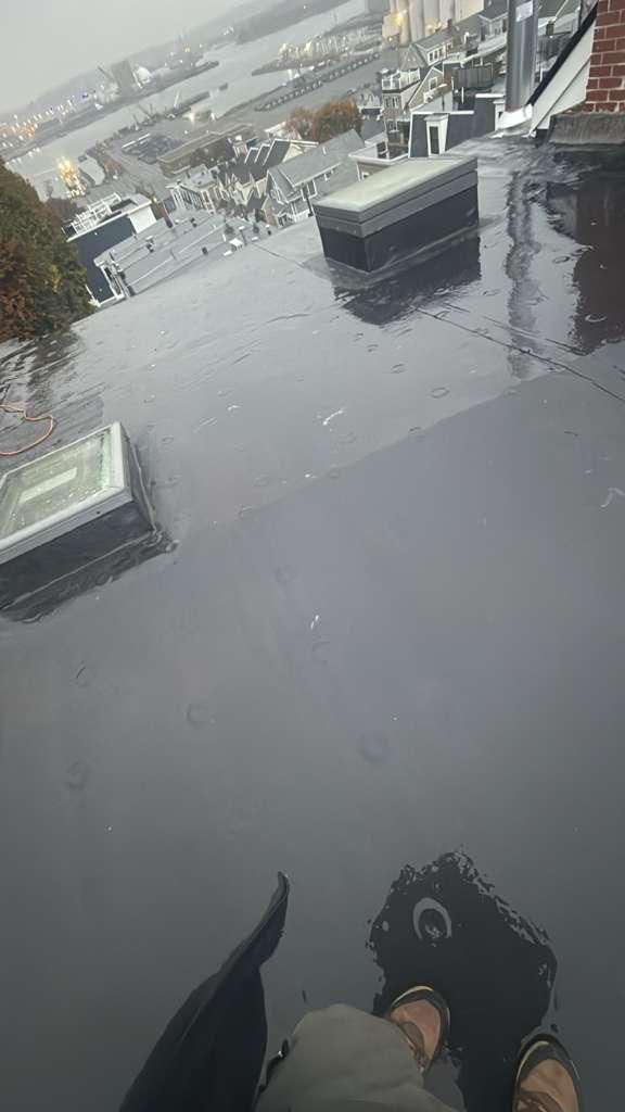 View from a wet rooftop overlooking a city. A person's feet and legs are visible, holding an umbrella.