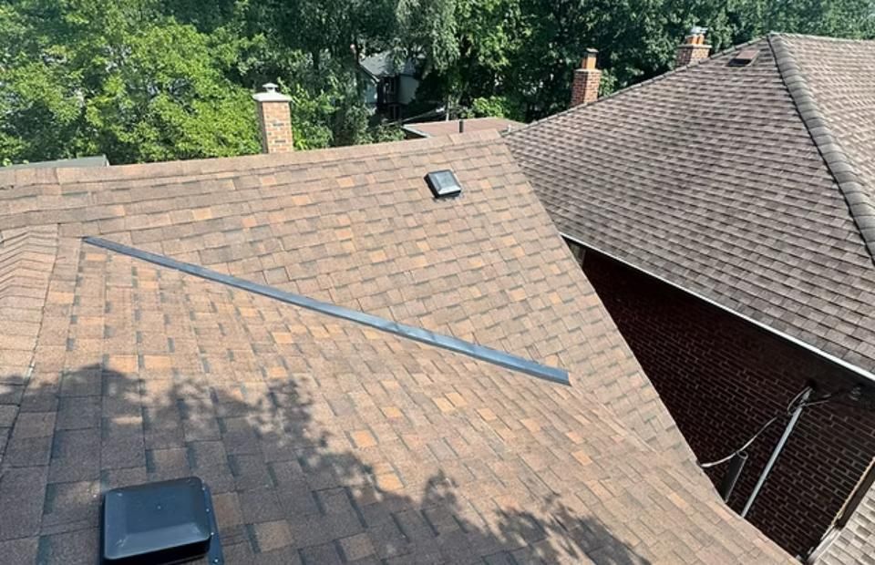 Brown shingled roof with a black metal strip, surrounded by trees and a brick wall.