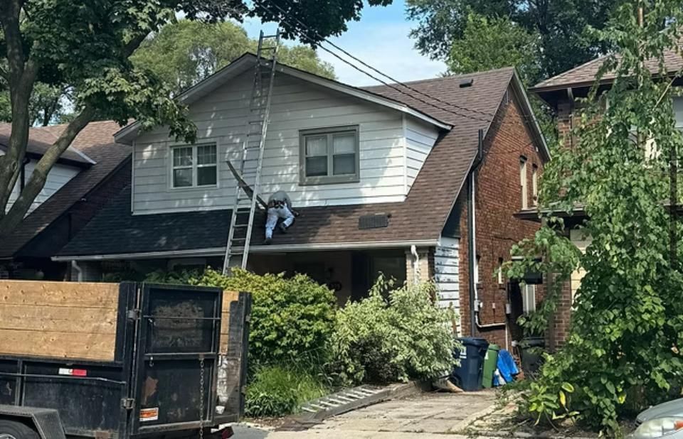 House undergoing repairs: worker on ladder, white siding, brown roof, dark truck in the foreground.