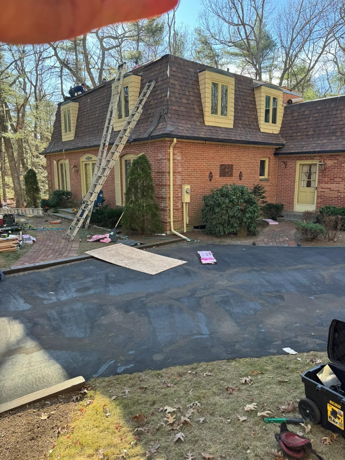 House with a ladder on the roof, roofers working. Asphalt driveway, trees, and tools visible.