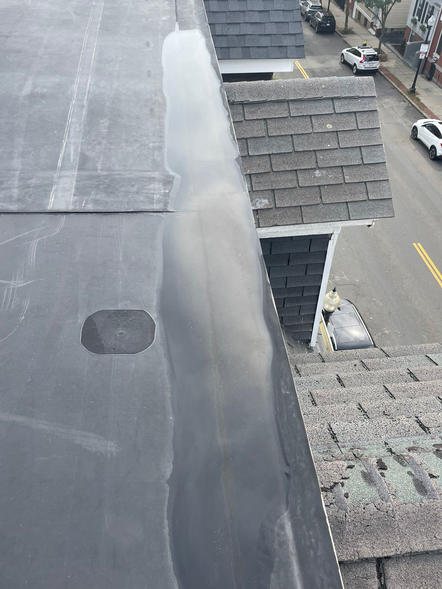 Flat, black roof with a long water puddle next to a shingled roof, street view in background.