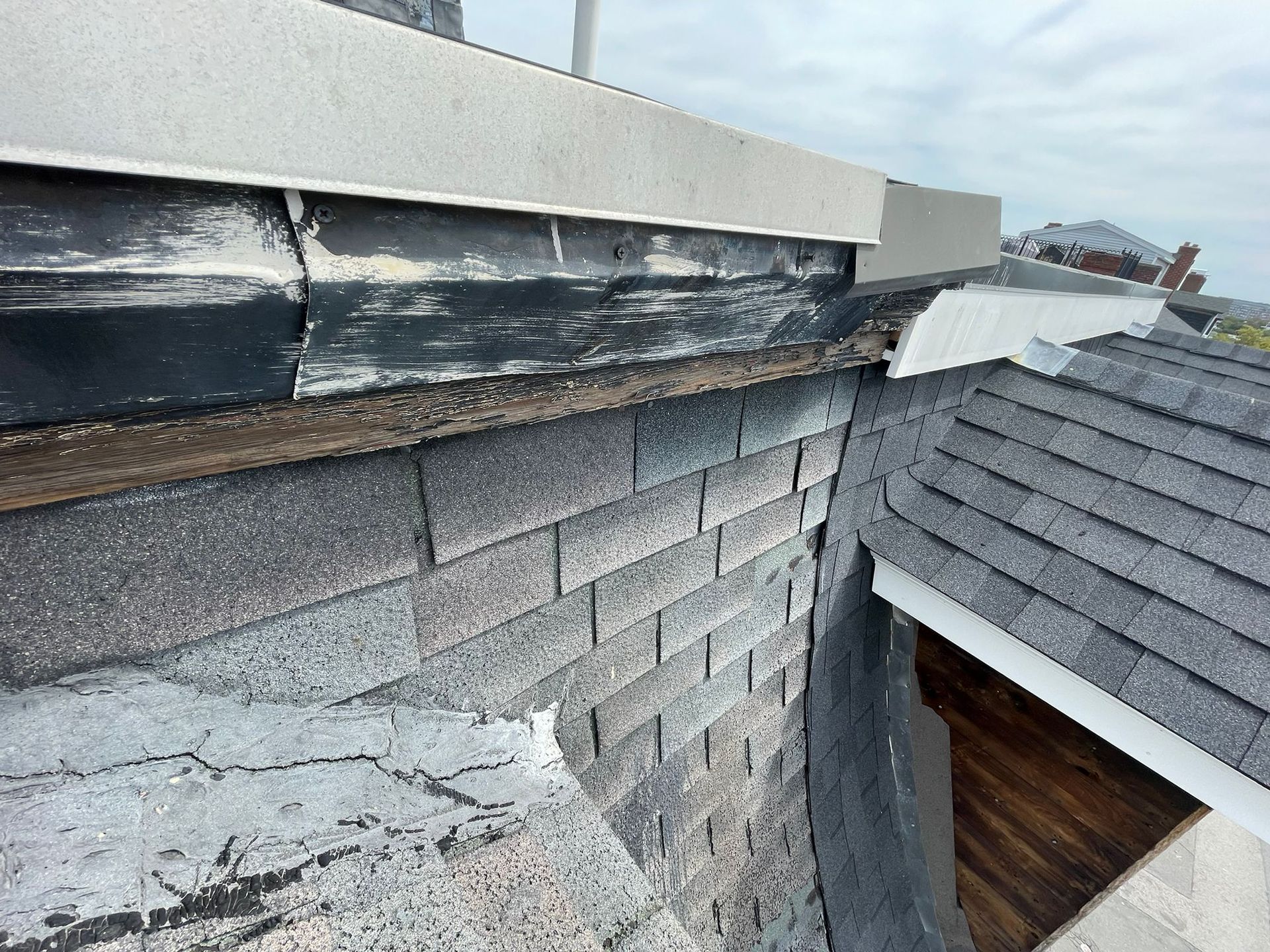 Close-up of a chimney and roof with damaged shingles and flashing. Gray and black hues dominate.