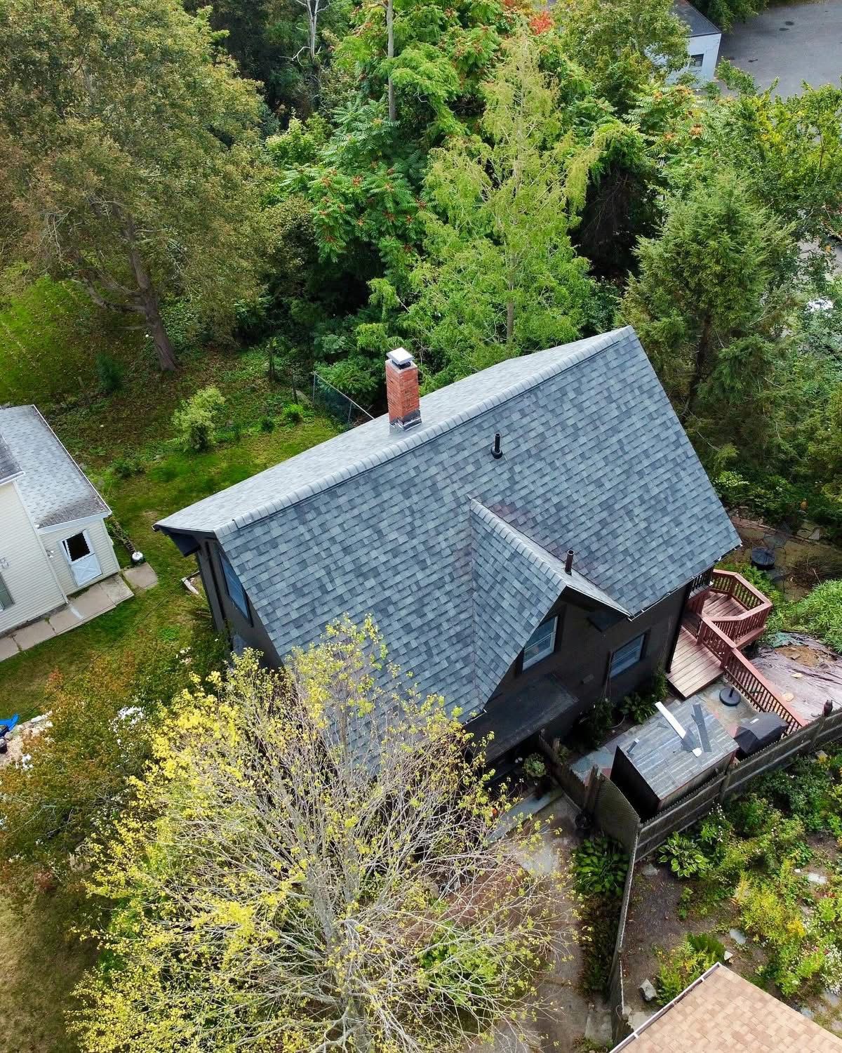 Aerial view of a dark house with a gray roof and brick chimney surrounded by trees and vegetation.