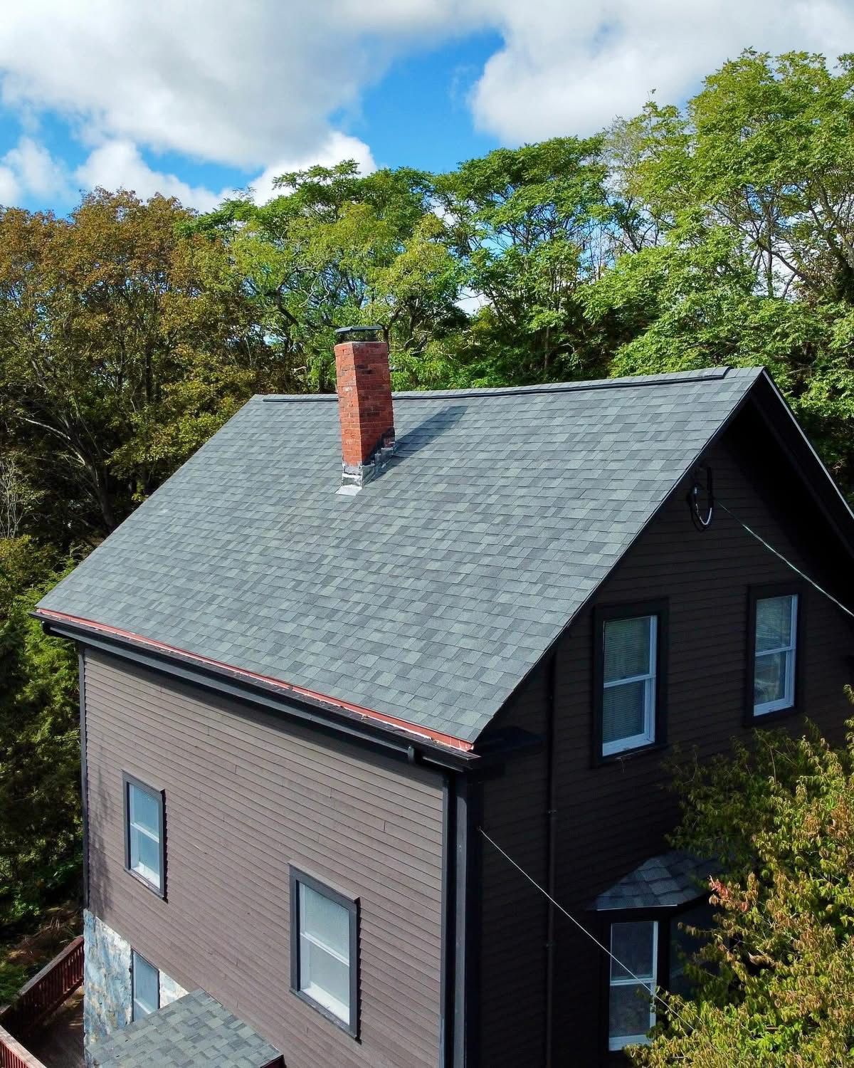 Roofer in yellow vest using a nail gun on a brown shingled roof under a cloudy sky.