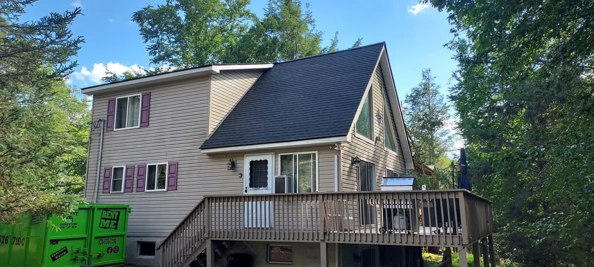 A-frame house with a deck and dark gray roof, surrounded by trees under a blue sky.