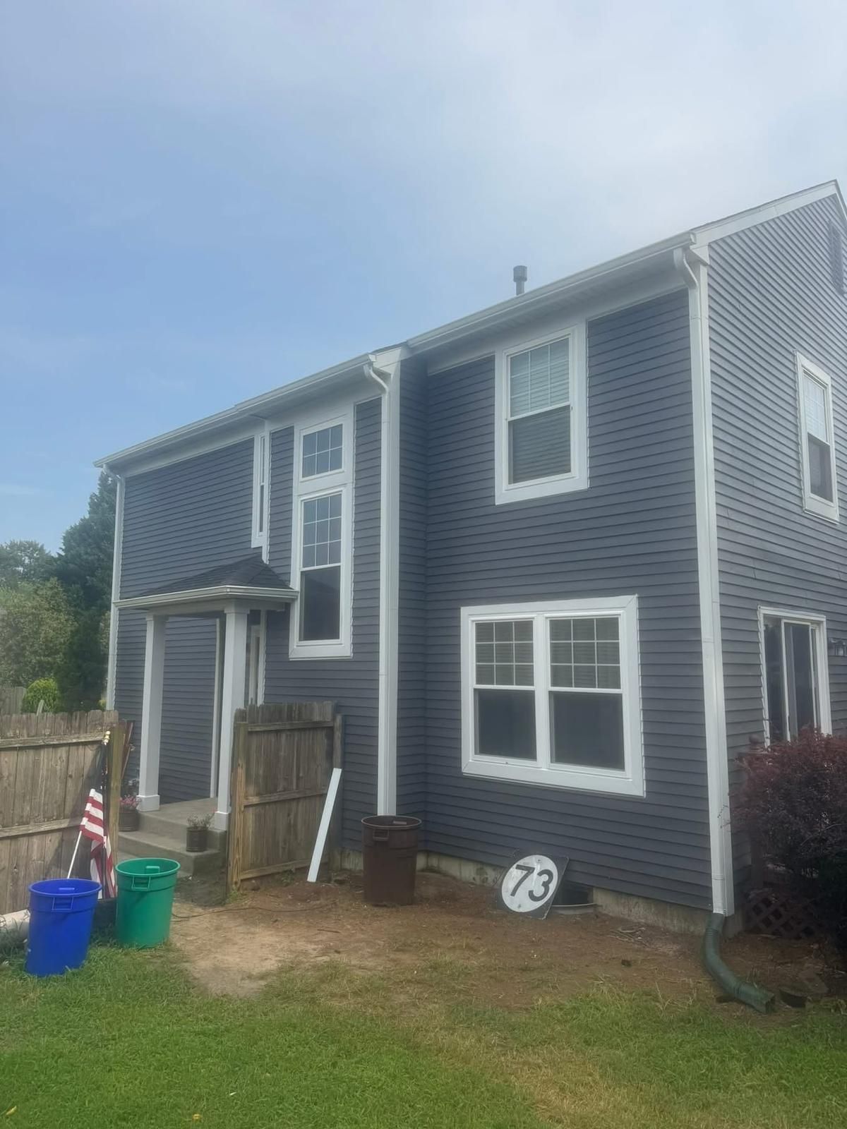 Two-story house with dark gray siding, white trim, and a small yard with debris.