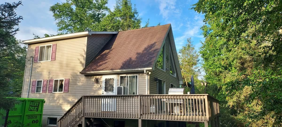 A-frame house with a wooden deck, surrounded by trees. The sky is blue.