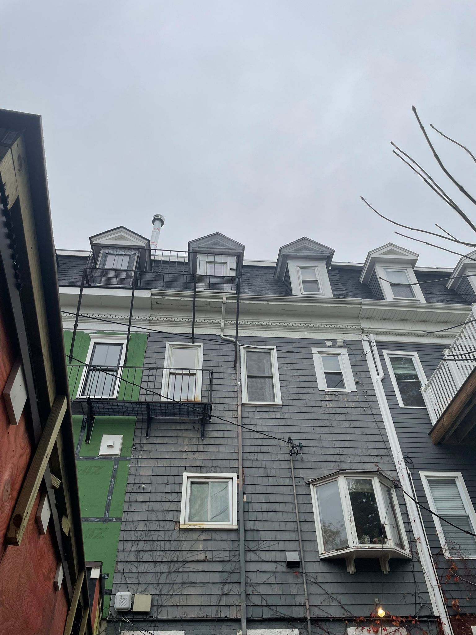 Gray and green building exterior with dormers, windows, and fire escape against a cloudy sky.