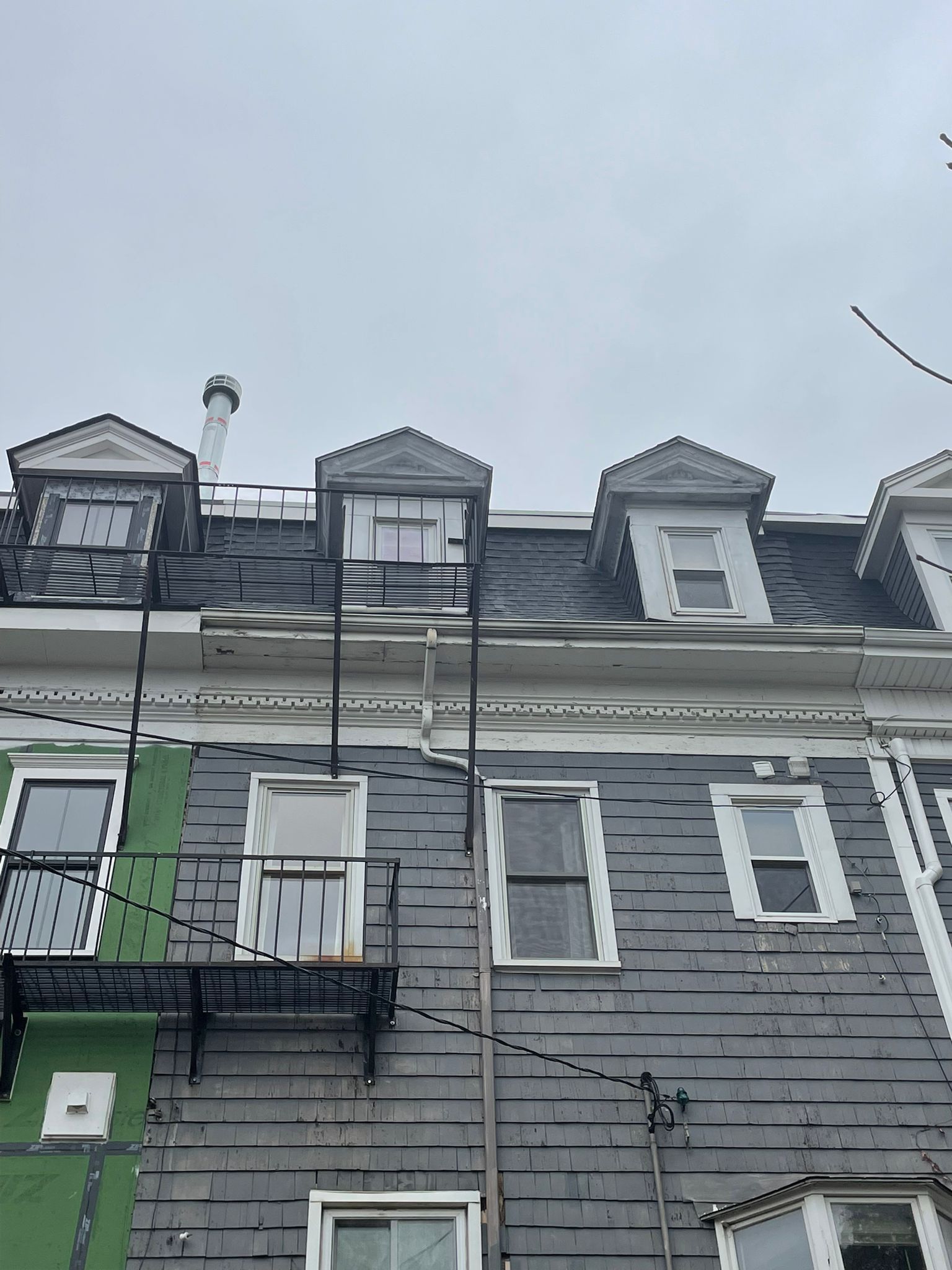 Row of gray and green weathered buildings with dormers and fire escape, cloudy sky.