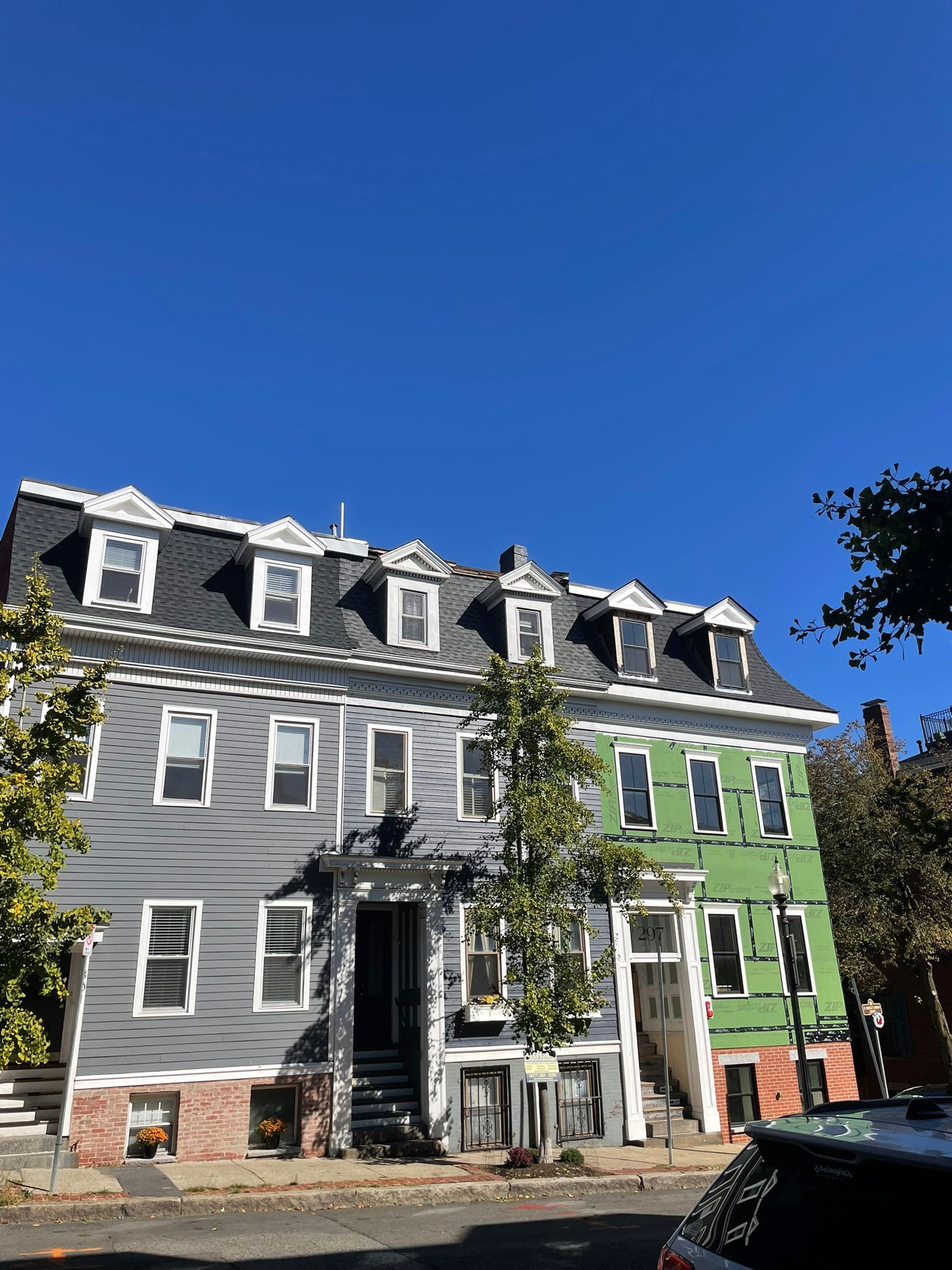 Row of multi-story townhouses, one gray, one light gray, one green, with dormer windows under a clear blue sky.