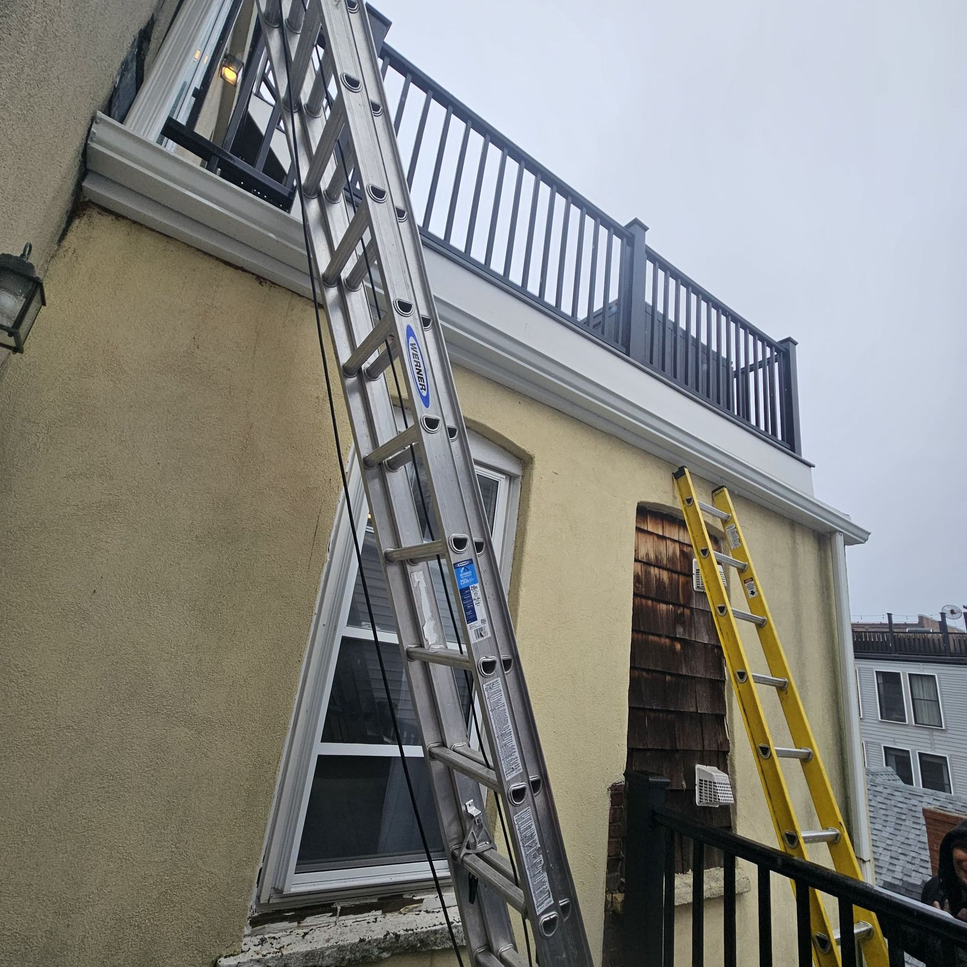 Two ladders leaning against a light-yellow building with a balcony. One is silver, the other yellow. Cloudy sky.