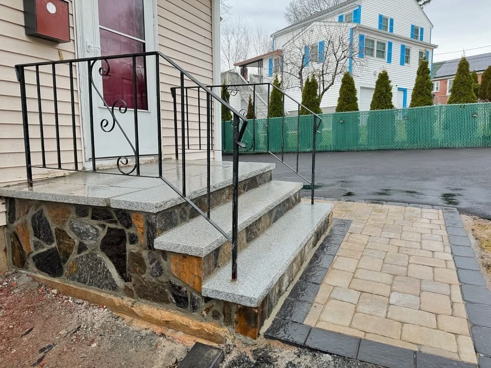 Stone steps with black railing leading to a house entrance. A brick walkway is to the right.