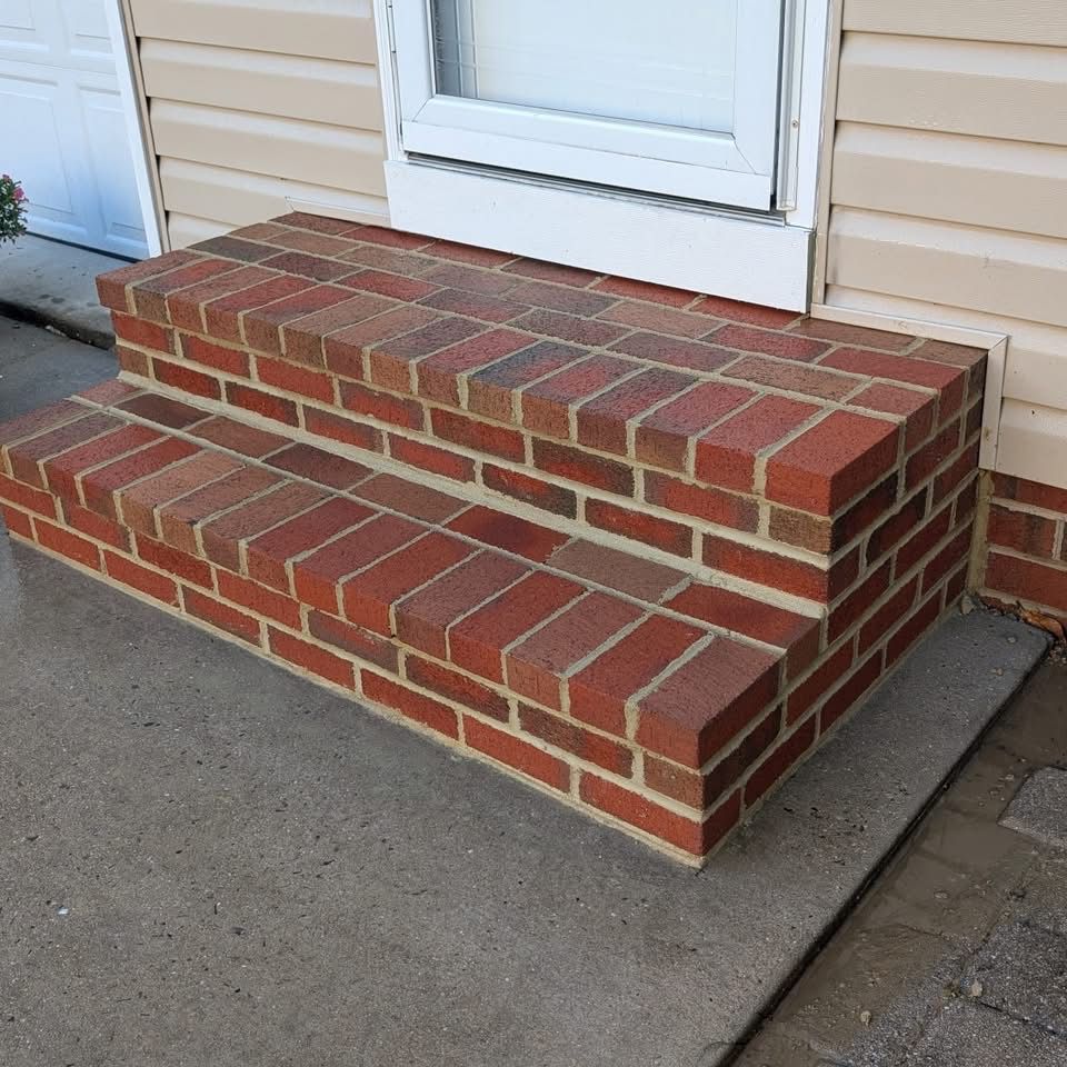 Brick steps leading up to a white-framed door on a beige siding house, set on a concrete slab.
