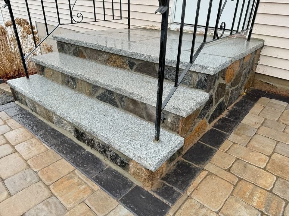 Granite steps with decorative stone facing and iron railing, surrounded by brick pavers.