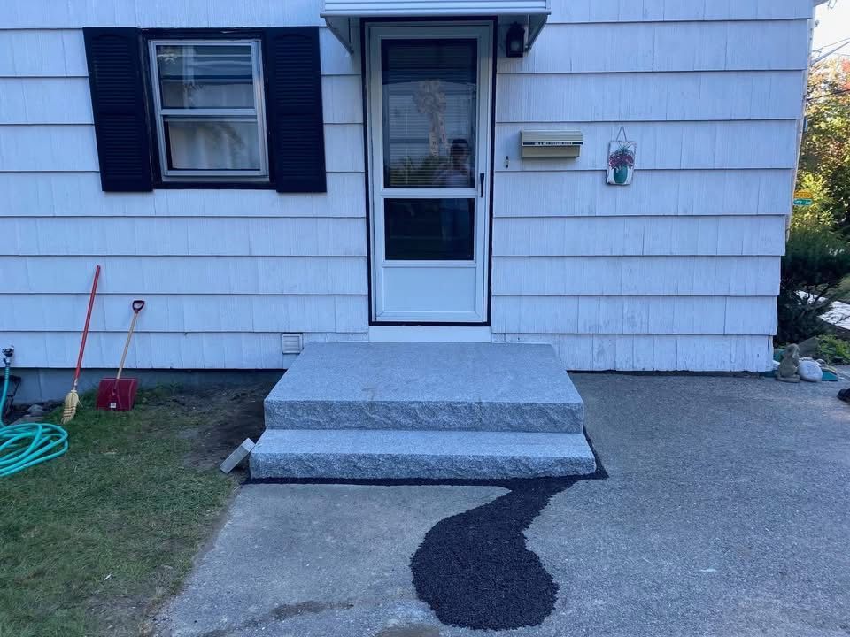 Exterior of a white house with granite steps leading to a door. A black spill sits on the concrete driveway.