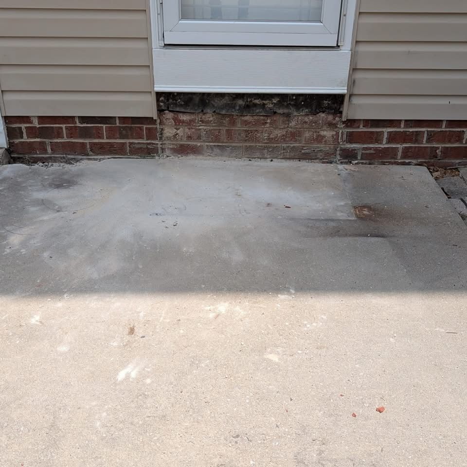 Exterior view of a building's base: beige siding, brickwork, a window above, and a concrete patio.
