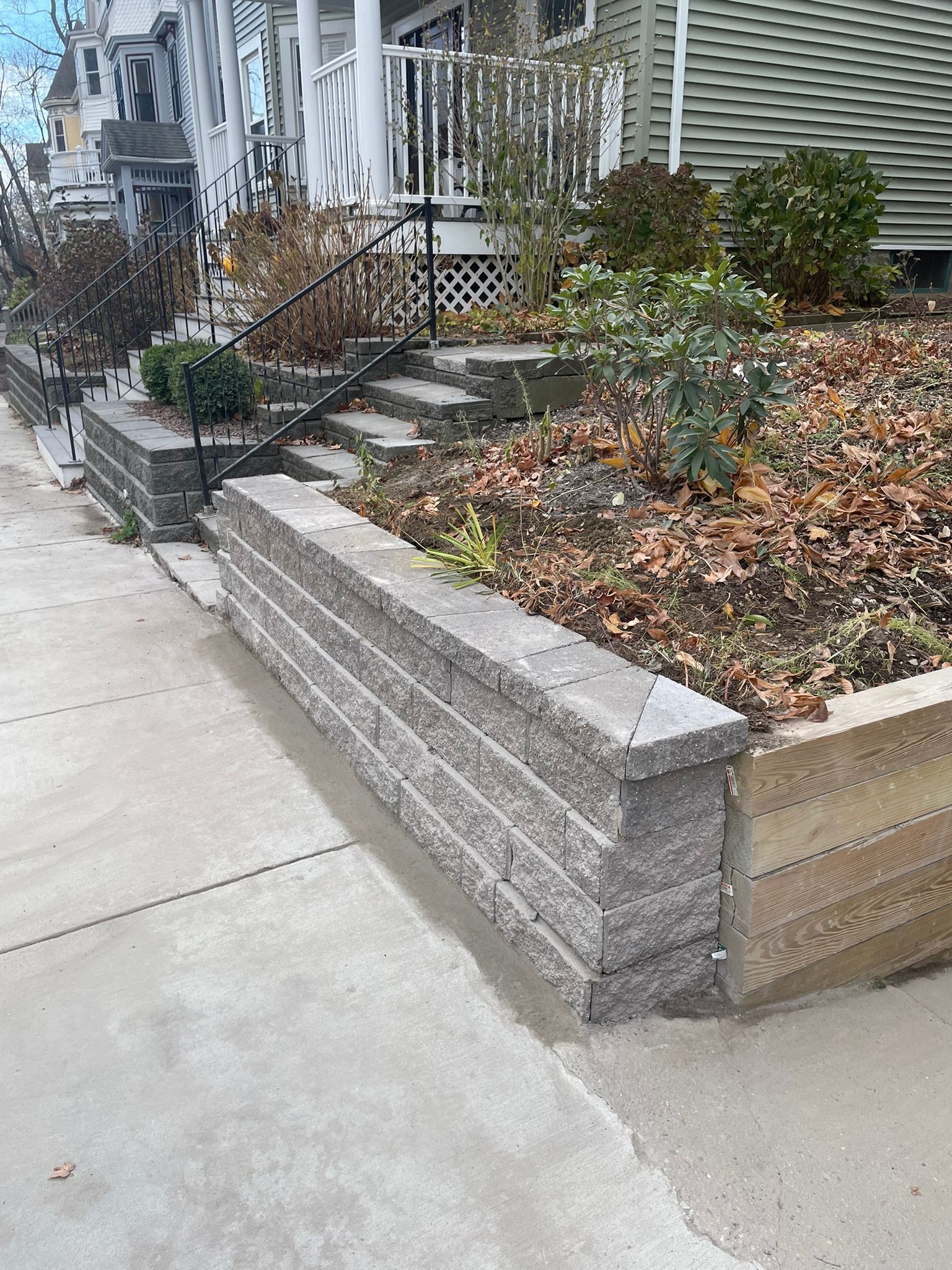 A gray brick retaining wall on a sidewalk in front of a house. Steps and plants are visible.
