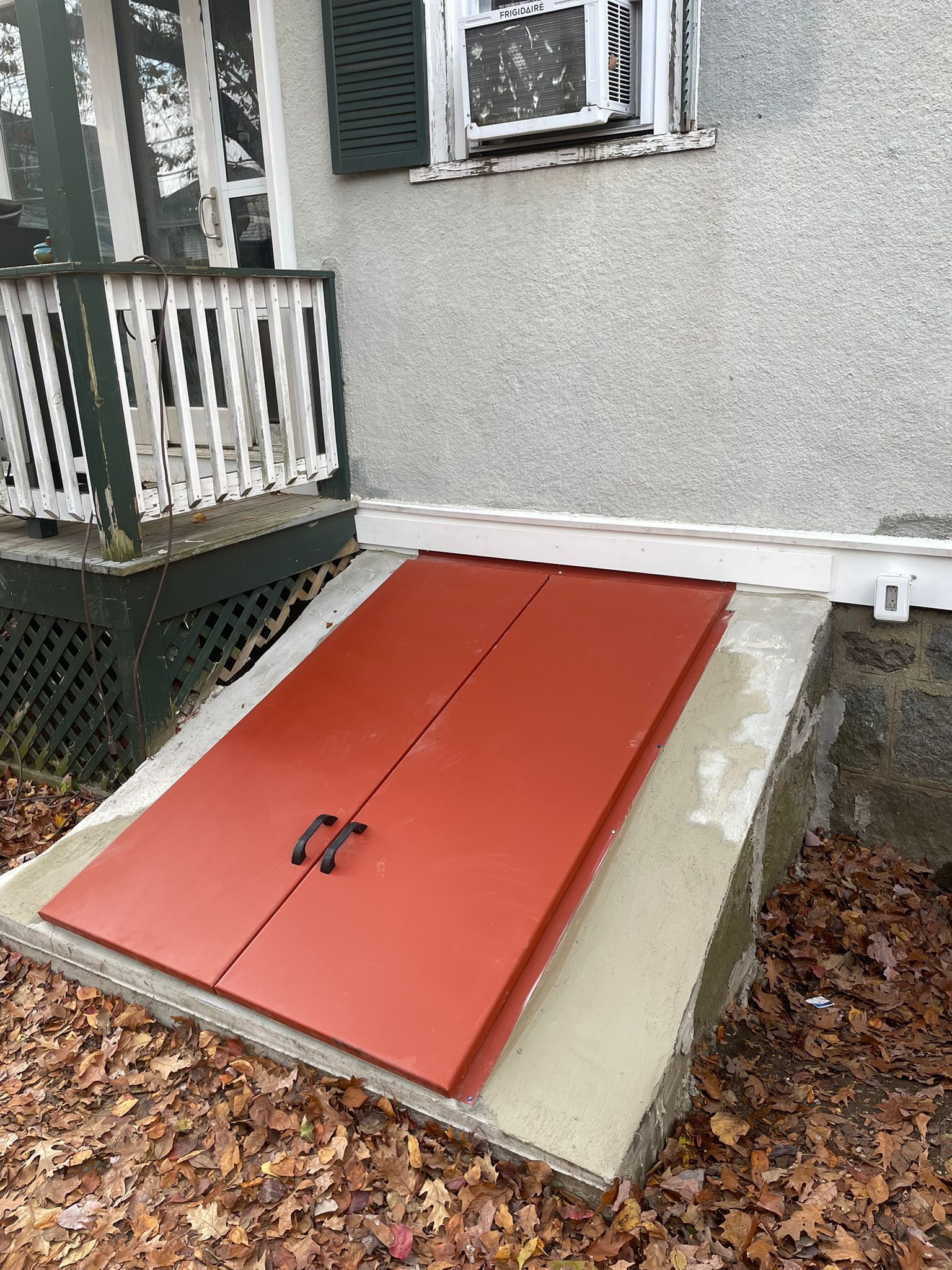 Red basement doors on a concrete platform, next to a house with a porch.