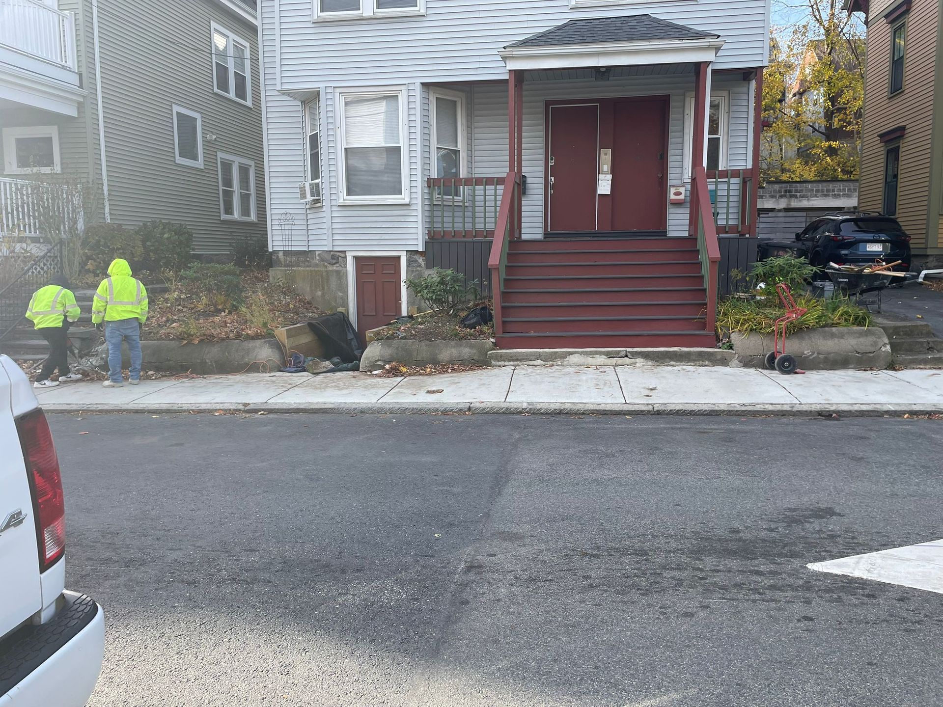 Two workers in reflective vests by a house with red door. Gray building, autumn leaves. White van foreground.