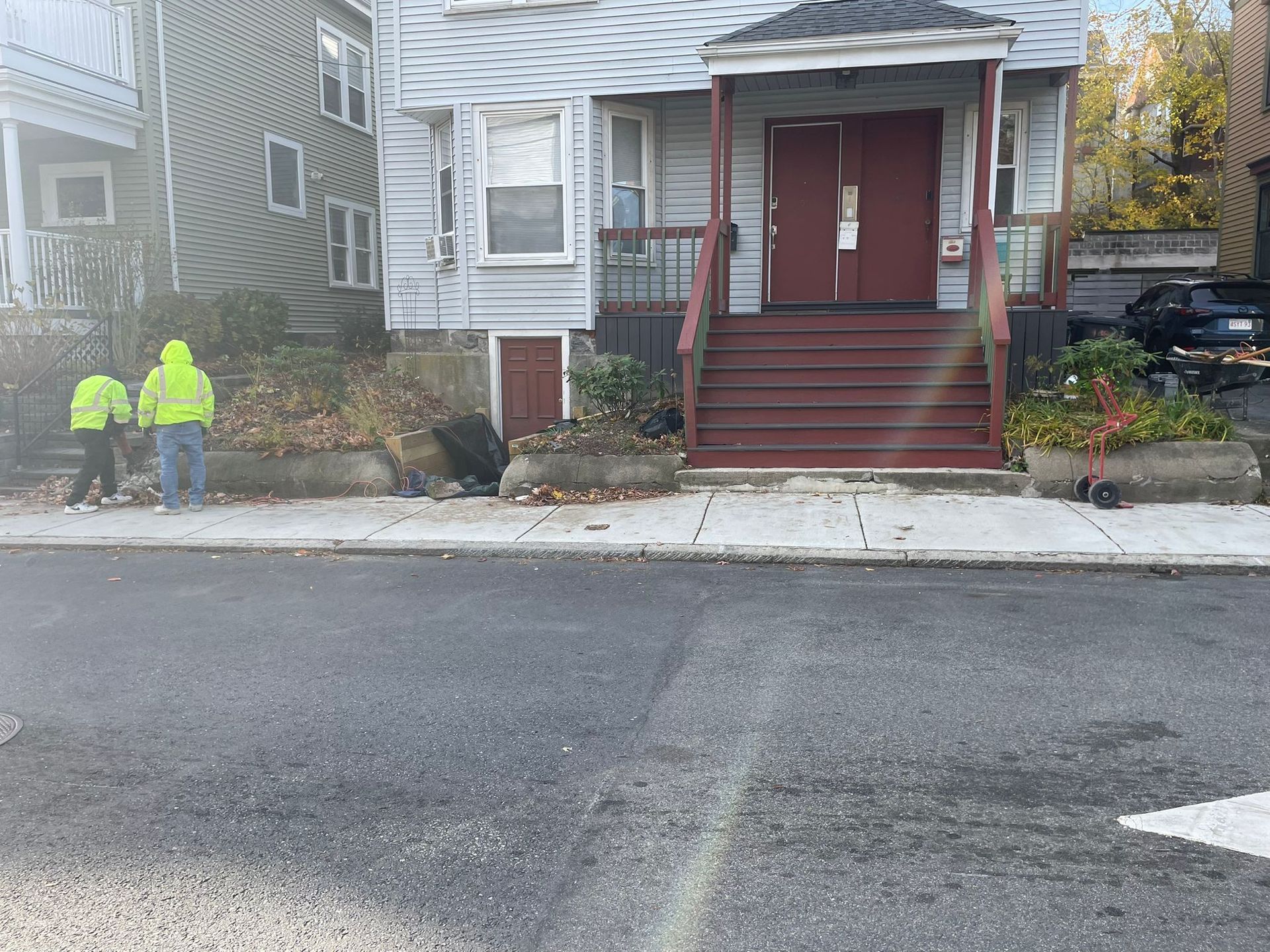 Two people in yellow vests clean debris along a sidewalk in front of a house with red doors.