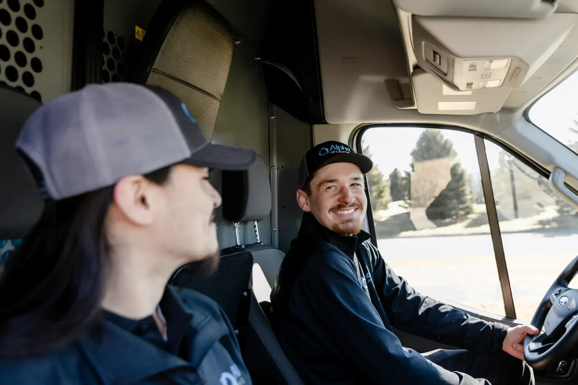 Two people in a truck cab, one driving and smiling, with sunlight through the windshield.