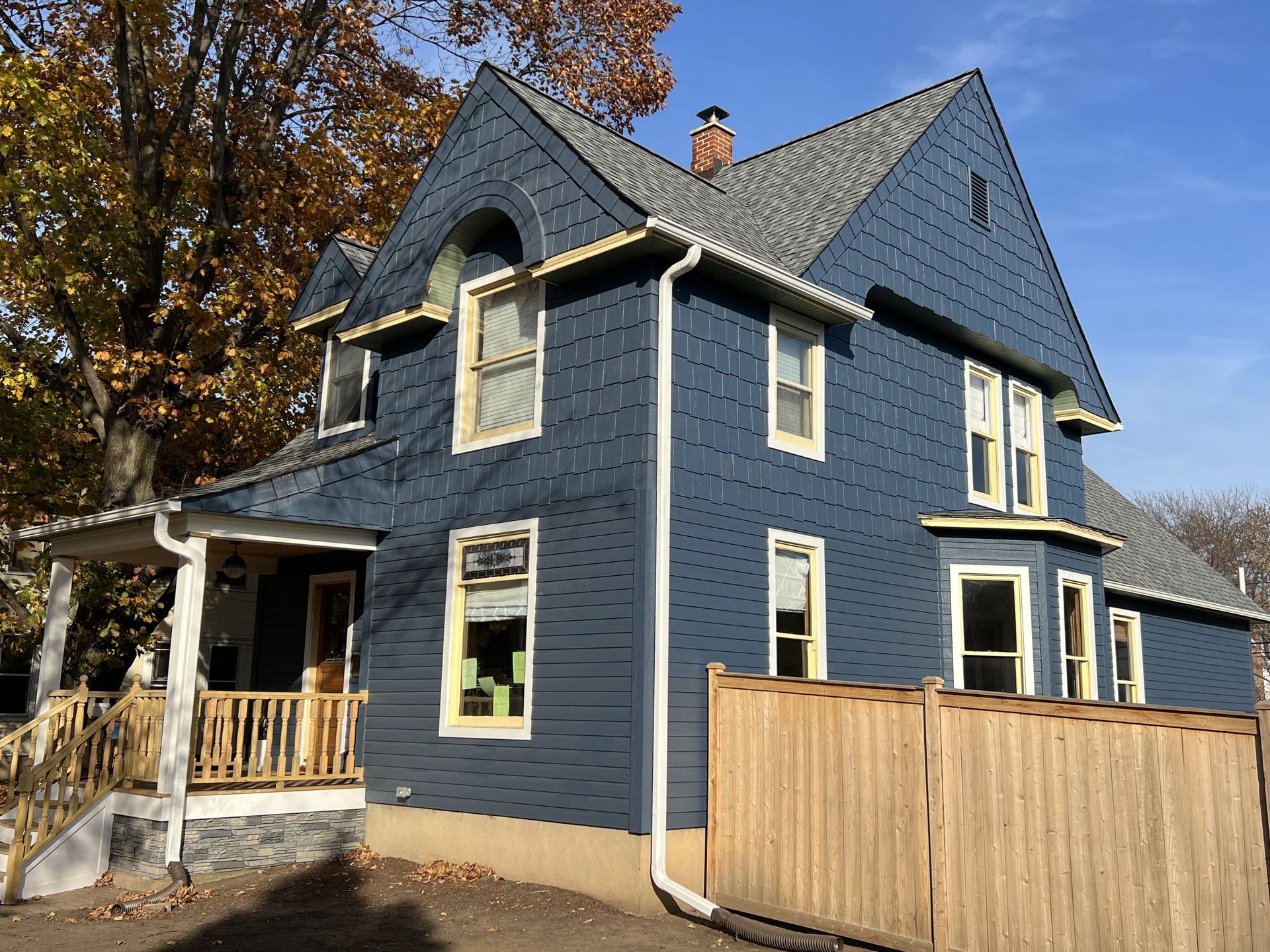 Blue two-story house with white trim, a porch, and a wooden fence in autumn.