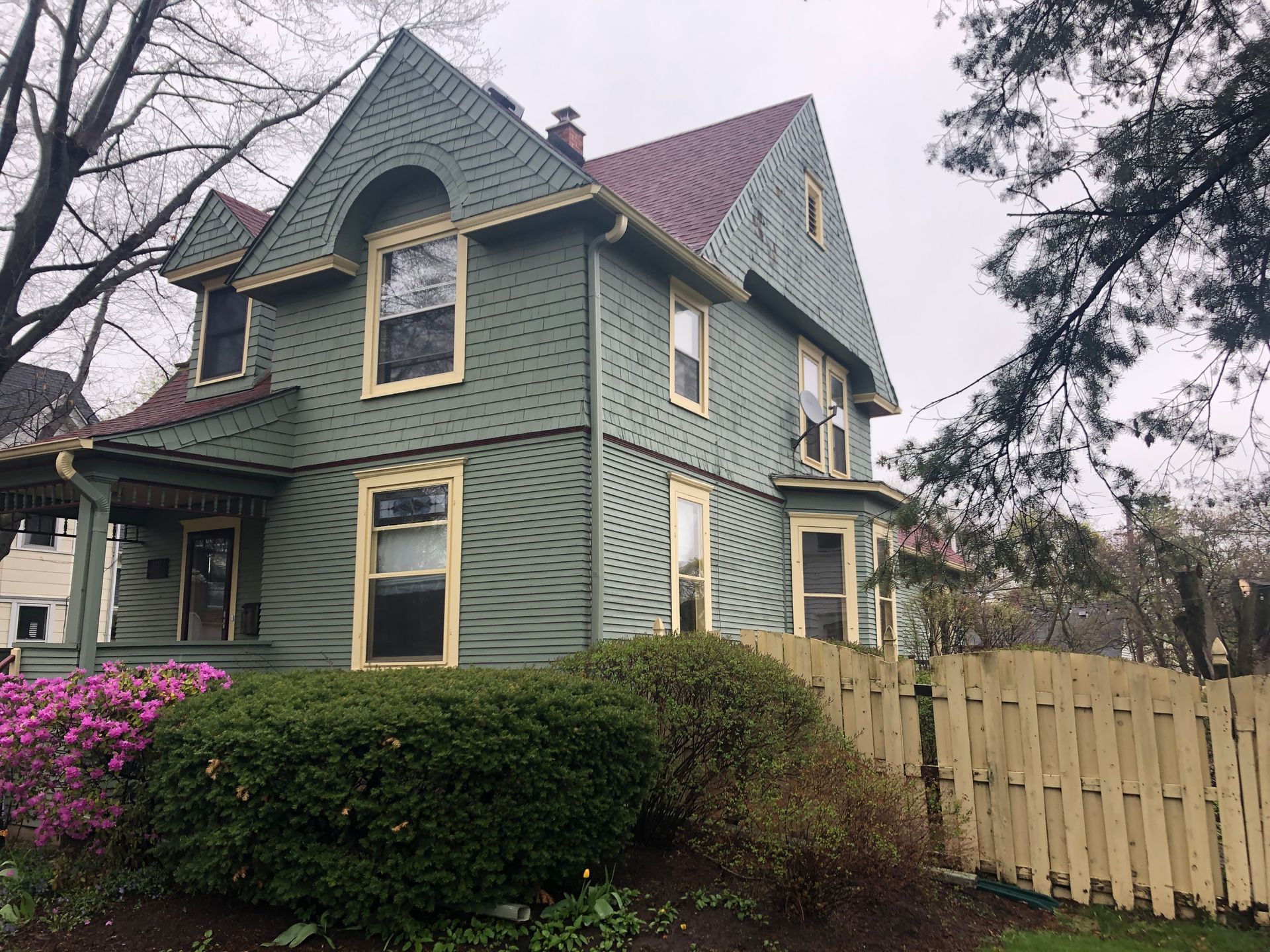 Green Victorian house with steep gables, white trim, and a wooden fence beside pink flowers.