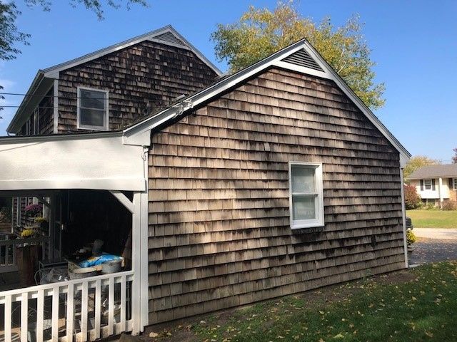 Weathered brown shingle house with white trim and a small porch under a blue sky