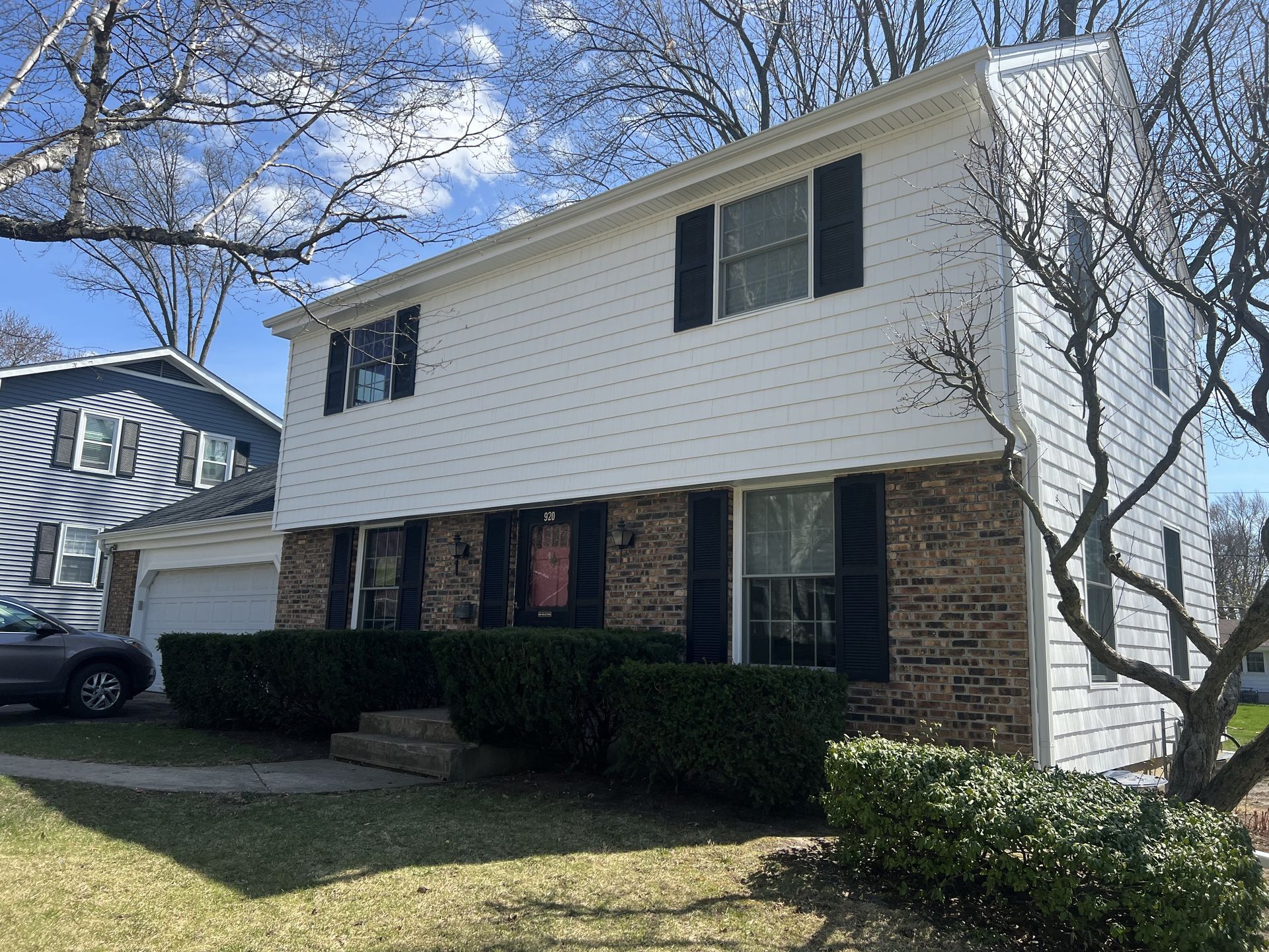 Two-story suburban house with white siding, brick lower level, black shutters, and trimmed hedges under a blue sky