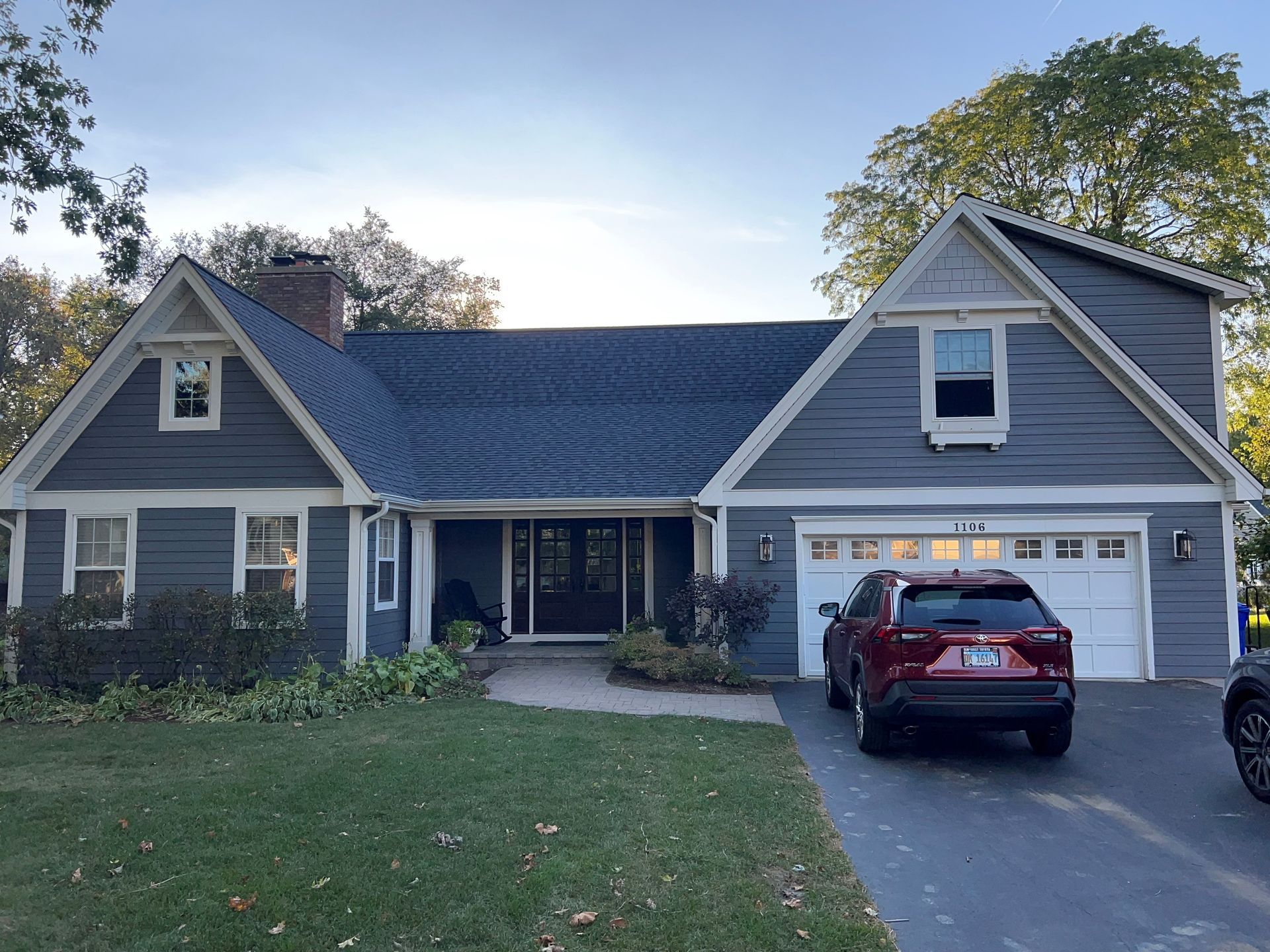 Suburban gray house with a front porch and garage, with two cars parked in the driveway at dusk
