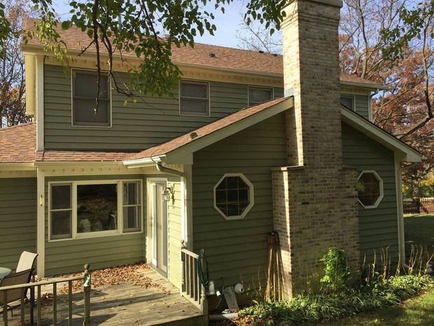Two-story green house with a stone chimney and front porch surrounded by trees.
