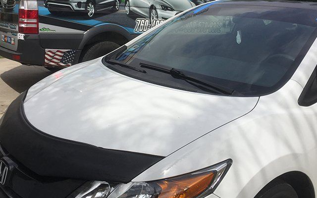 A white car with a black hood protector is parked on the side of the road.