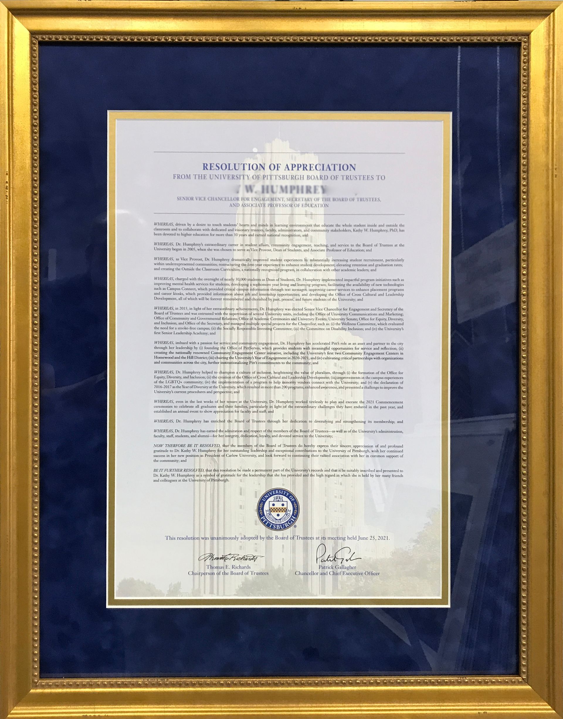 Framed document, likely a certificate, set against a blue background within a gold frame. Official seal at the bottom.