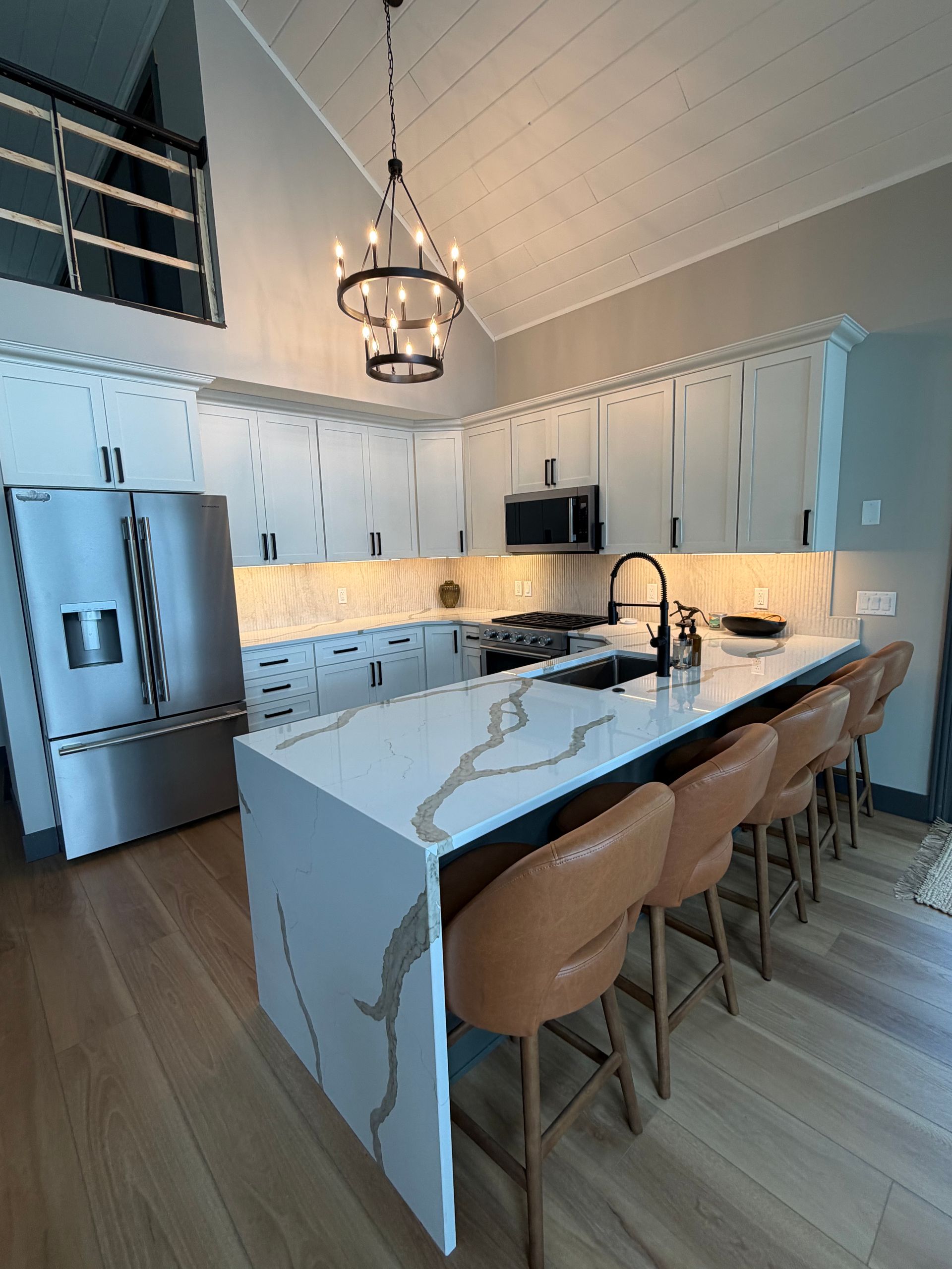 Modern white kitchen with marble island, tan bar stools, stainless fridge, and pendant light