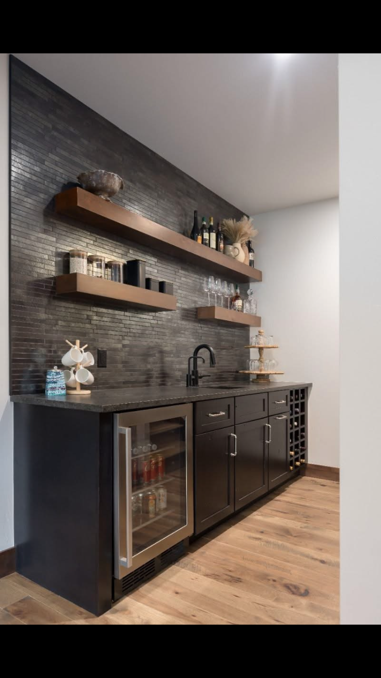 Dark home bar with black cabinets, stone backsplash, wooden shelves, and mini-fridge.