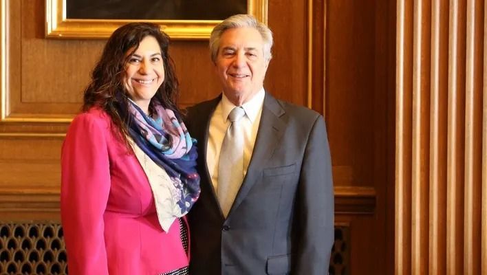 Two people standing indoors, smiling, in formal attire against wood-paneled walls.
