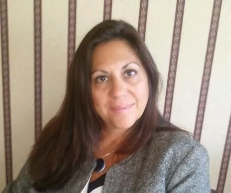 Portrait of a woman with long dark hair, wearing a gray cardigan, standing against striped wallpaper.