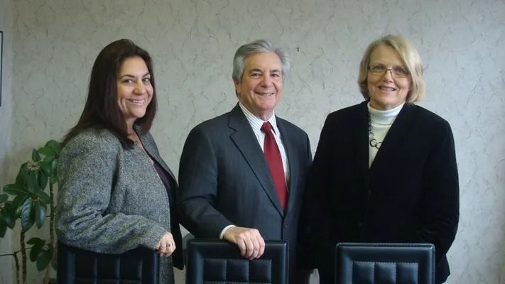 Three people stand indoors, smiling behind chairs, with a man in a suit between two women in dark coats.
