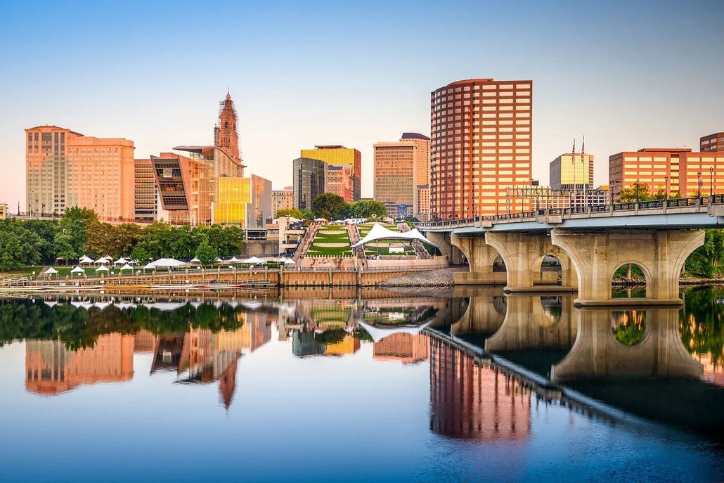 City skyline at sunset reflected in a calm river beside a bridge.