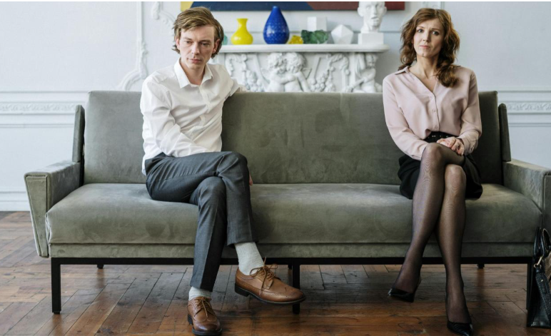 Two people sitting apart on a green sofa in a bright living room, looking somber.