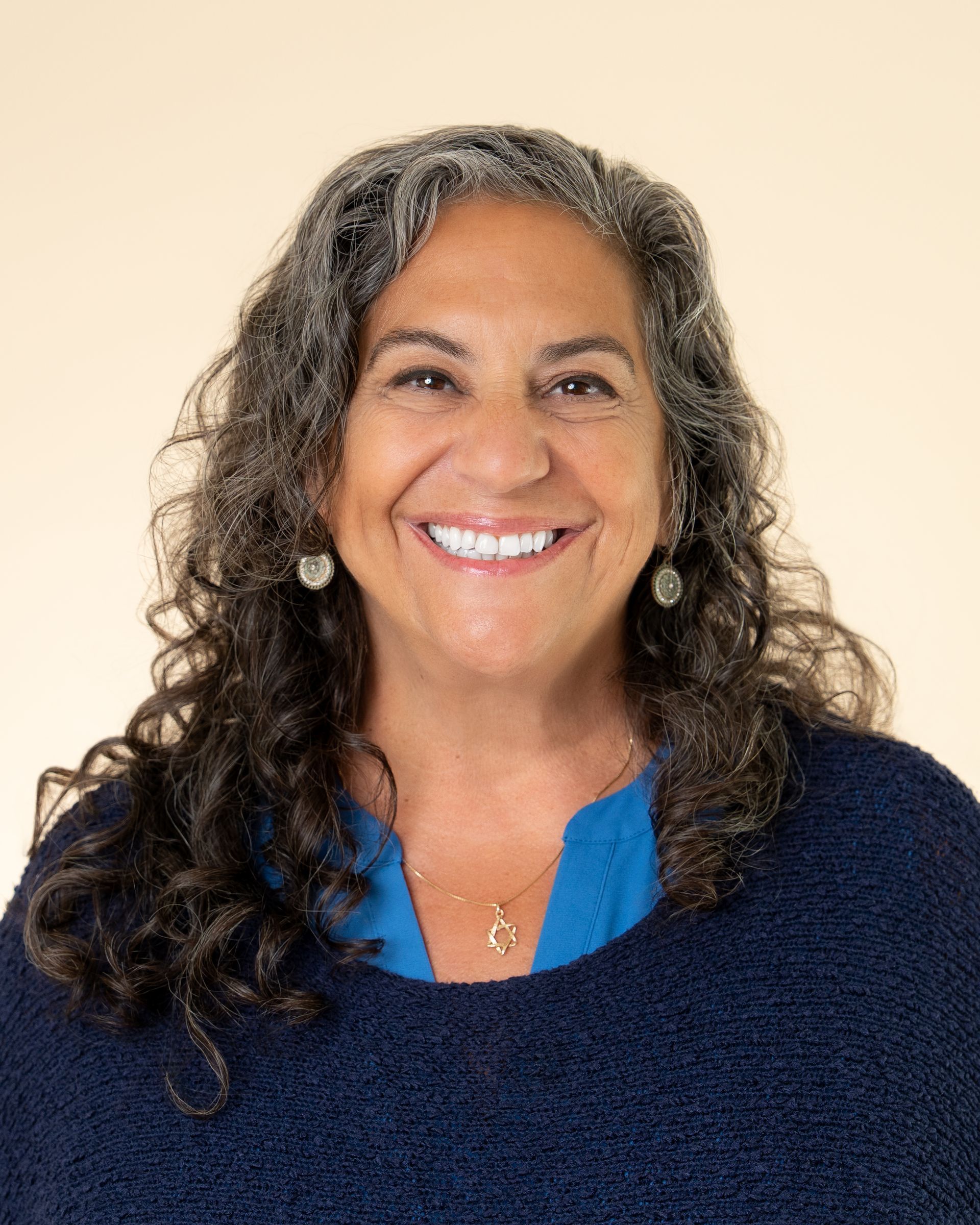 Smiling woman with curly gray hair wearing a navy sweater and blue top against a beige background