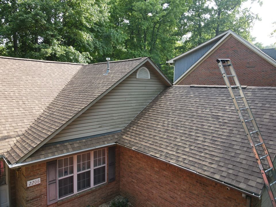 Brown-shingled house exterior with a ladder against the brick wall, surrounded by trees.