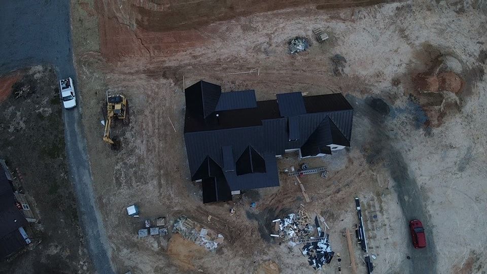 Aerial view of a house under construction with a black roof, surrounded by dirt and construction equipment.
